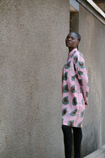 Model standing against a textured wall, wearing the pink Press Dress with green and black avocado print, paired with black leggings.