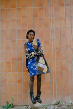 The Press Dress in Virtual Reality batik print, featuring a mix of blue, white, and yellow patterns, styled with black boots, against an orange tiled wall.