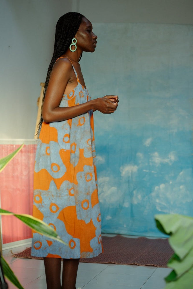 Serene indoor vibe with Saya Dress in orange and blue floral pattern, blue sky wall, pink canvas, and green plant.