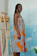 Side view of the Saya Dress in blue and orange All Ideas print, artistic setting with ladder and woven basket.