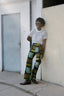 Veronica Trousers in Pistacia batik print, worn with a white t-shirt, standing against a white wall next to a gray door.