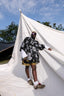 Patterned Album Garment top paired with dark shorts and light brown shoes, standing beside a white tent under clear skies.