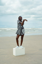 A person wearing a black and white patterned dress stands on a white box at the beach.