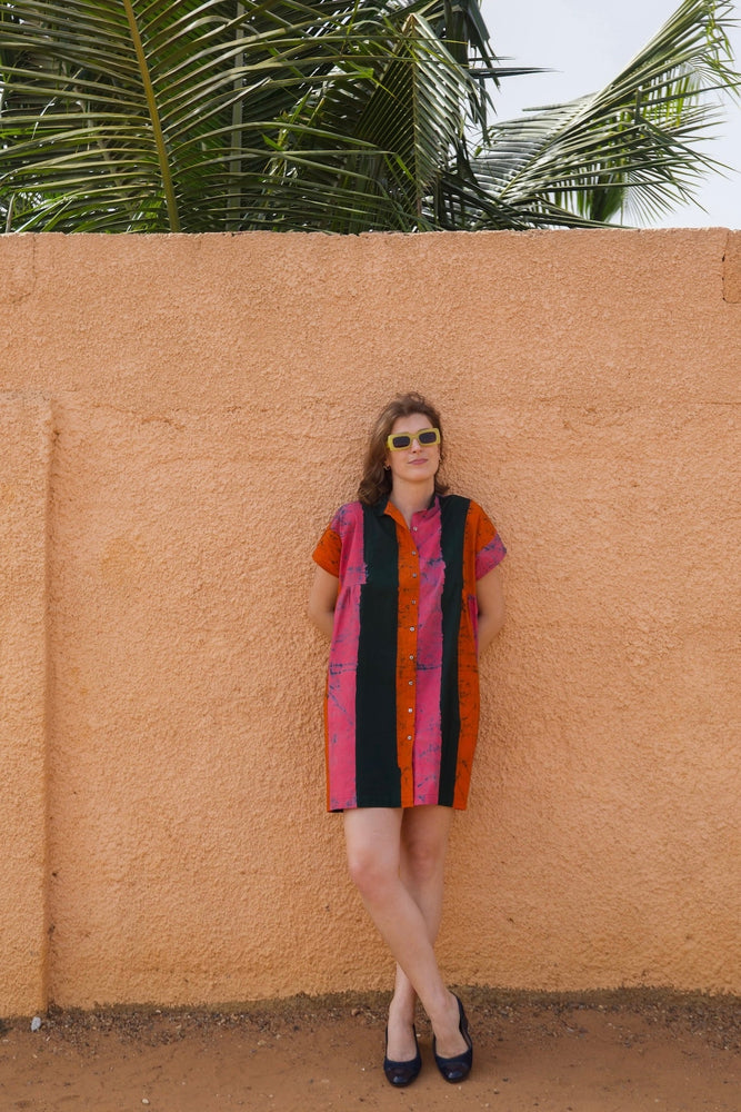 Colorful Bata Dress in Carmine print against an orange wall, complemented by black flats and tropical greenery.