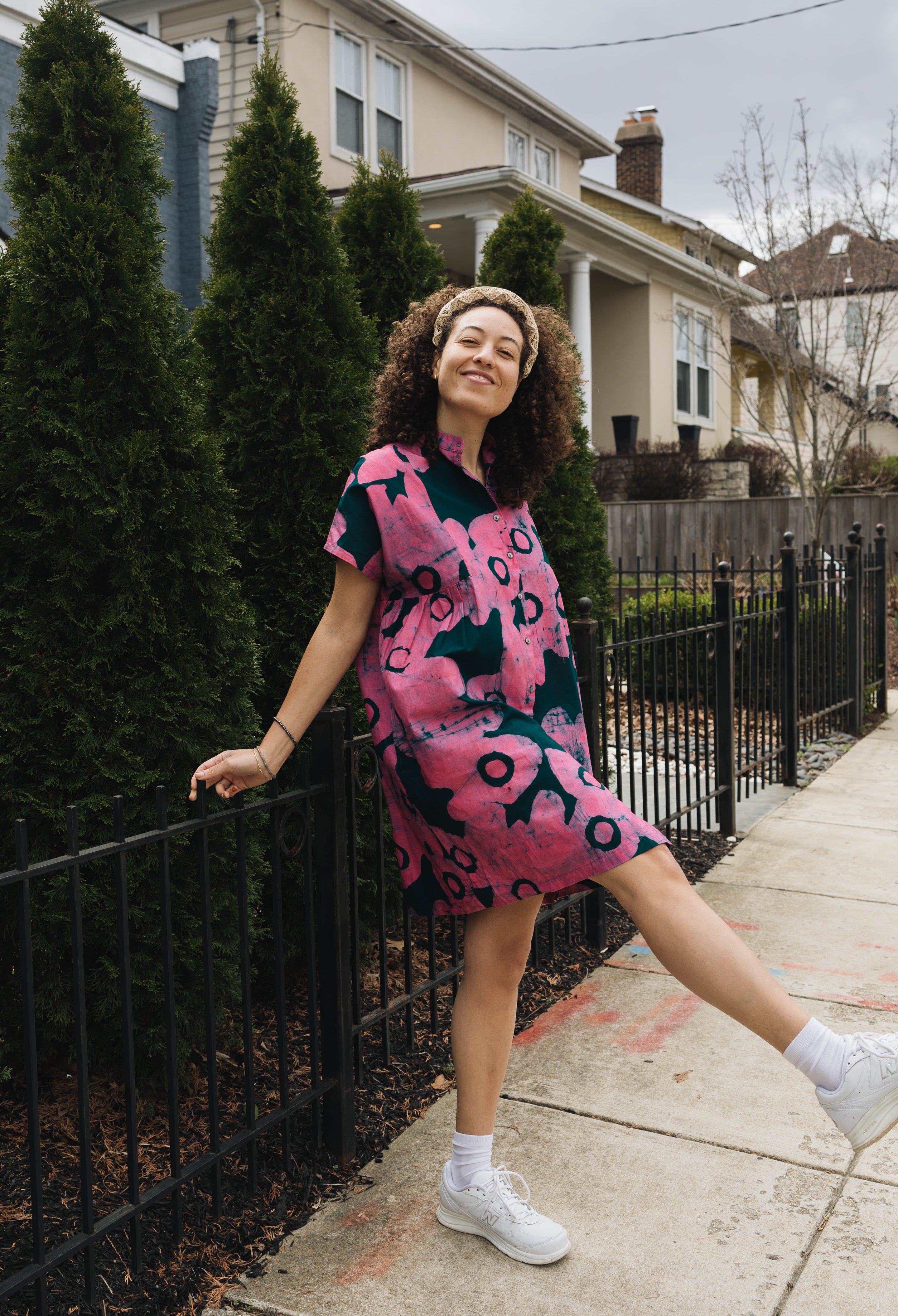 A smiling figure wearing a knee-length black and red patterned dress leans on a black fence while raising one leg up slightly.