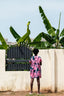 Rear view of Bata Dress in pink with blue floral pattern, against a white wall with tropical banana leaves backdrop.