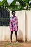 Colorful Bata Dress with abstract floral print, white sandals, in front of white wall with metal fence and green plants.