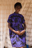 Person in blueish- purple Bata Dress, abstract pattern, hands clasped, standing before patterned curtain.