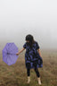 Serene Bata Dress in foggy field, purple umbrella in hand, showcasing unique print and style.