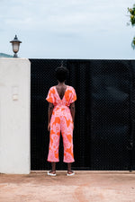 Easy Jumpsuit in Tee Hee at a black gate, orange with pink print, outdoor lantern on white wall, cloudy sky.