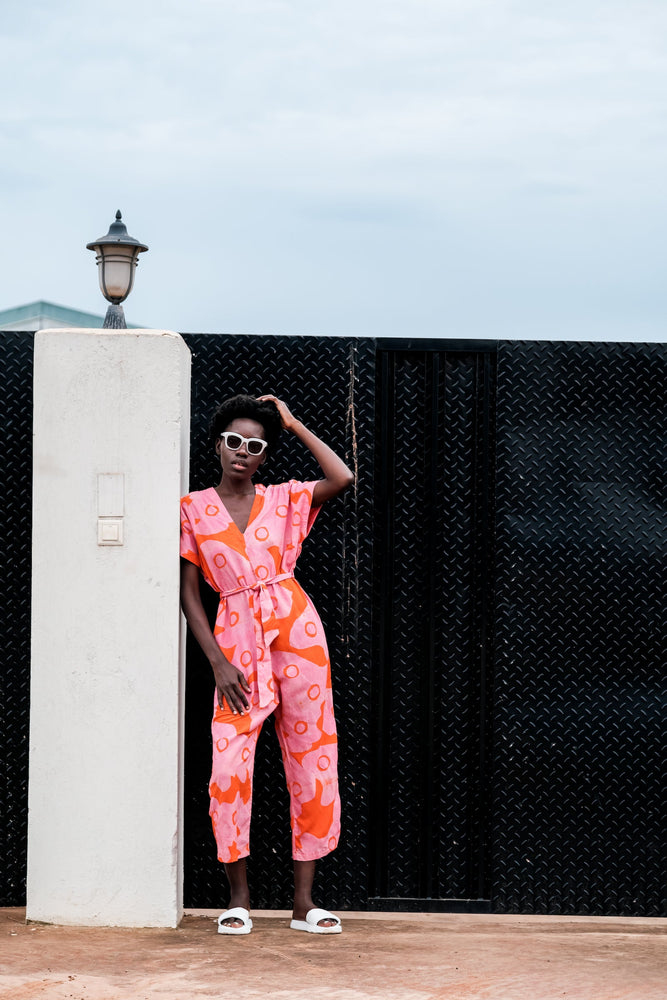Easy Jumpsuit in Tee Hee beside a white pillar and lamp, featuring pink and orange batik print, against a black gate.