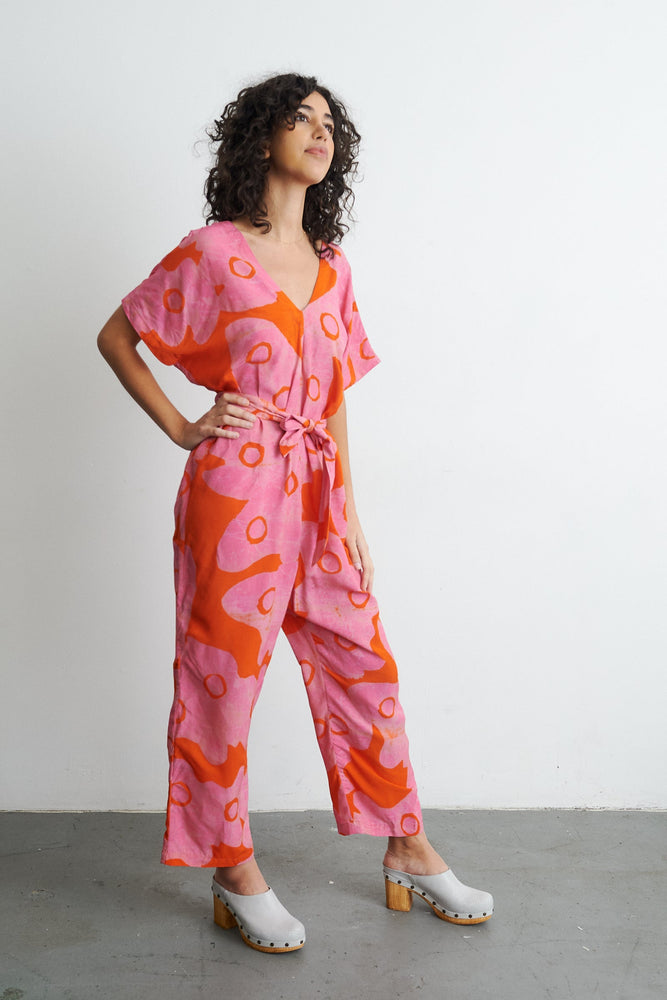 Person in Easy Jumpsuit with vibrant pink and orange pattern, posing with hand on hip, in clogs against white wall.