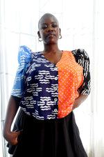 Colorful, multi-section blouse with unique patterns. Model stands against a sheer curtain backdrop. Ethically made in Ghana.