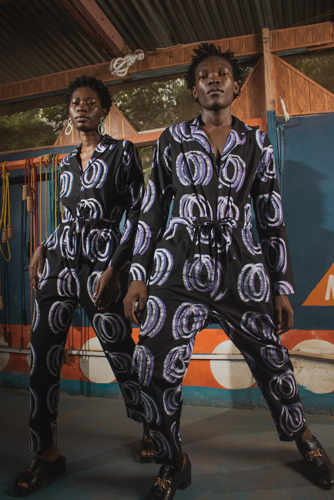 A man and a woman in matching Accra Jumpsuits with Good Signal print, posing in a colorful interior setting.