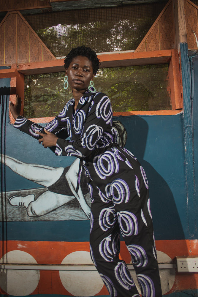 Fashionable Accra Jumpsuit in Good Signal print, black with white and purple patterns, in front of a vibrant mural.