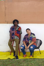 Our rayon unisex Holiday Shirt in Hocus Pocus print worn by a man and a woman sitting on a chair in front of a wall.