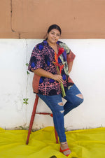 A woman sitting on a stool with flowers, wearing our colorful rayon Holiday Shirt in the 3 way split print, Hocus Pocus.