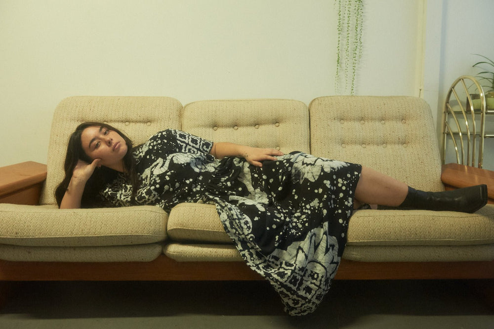 Relaxed Imperium Dress in 2 Party System print, styled with black boots, lounging on a mid-century sofa with chic decor.