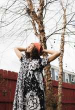 Chic Imperium Dress in monochrome floral, paired with a red hat, against a rustic red fence and bare tree backdrop.