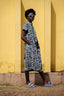 Chic Imperium Dress in 2 Party System print, paired with light blue sandals, against a textured yellow wall.