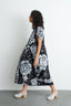 Dynamic Imperium Dress in 2 Party System print, mid-step movement, paired with white clogs, against a minimalist backdrop.