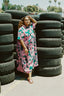Imperium Dress amidst stacks of used tires, floral pattern standing out against an outdoor backdrop with clear skies.
