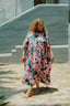 Imperium Dress in lively Pool Party print, outdoor setting with textured blue wall, embodying casual elegance.