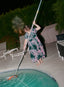 Person skimming pool at night, wearing a floral dress, beside an illuminated pool with lounge chairs and lush greenery.