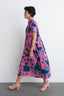 Imperium Dress in Pool Party print, mid-step in pink shoes, against a stark white backdrop, exuding dynamic elegance.