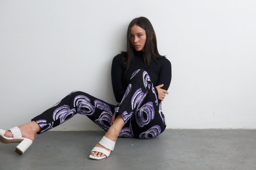 Kpong Trousers in Good Signal print, black top, white heels, sitting casually, embodying relaxed fashion.
