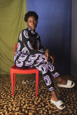 Kpong Trousers in Good Signal print, full-sleeve top, white sandals, seated on red chair, against fabric backdrop.