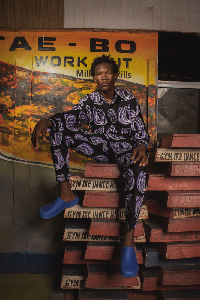 Kpong Trousers in Good Signal print, casual pose on wooden steps, vibrant urban setting, hand-dyed in Ghana.