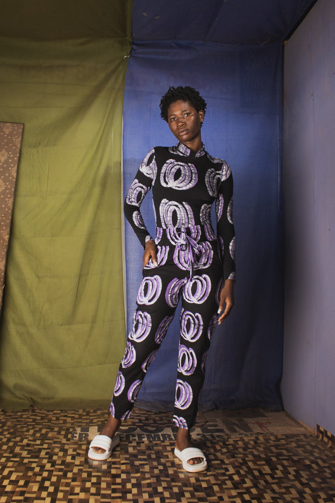 Kpong Trousers in Good Signal print, full-sleeve top, white sandals, against vibrant fabric backdrop.