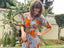 A woman wearing a beautiful orange and blue rayon wrap dress stands gracefully beside a prickly cactus.