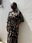 A woman in a black & tan print dress with batwing sleeves and a deep V neckline, leaning against a wall.