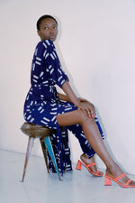 Letsa Dress in Middle Path print, seated pose highlighting side slits and wrap waist, with coral heels and rustic stool.