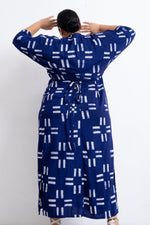Rear view of Letsa Dress in Middle Path print, blue with white line patterns, arms raised, with a wrap-around waist tie.