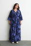 Blueish purple and black Letsa dress with V-neck and waist tie, wide sleeves, against white wall on grey floor.
