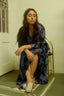 Person seated on golden chair in a blueish purple and black Letsa Dress, white sandals, with night view through window.