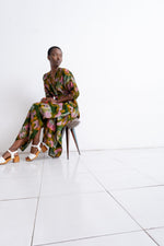 Colorful Letsa dress with green, yellow, pink patterns and white sandals, modeled sitting on a simple chair.