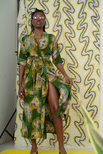 Model in Letsa dress with colorful abstract shapes, lifting hem to reveal leg against yellow-white backdrop.