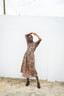 Nonna Dress in Snakebite print, model poses with arms raised, black shoes, against white wall and clear sky.