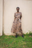 Nonna Dress in Snakebite print, black spots on a camel, against beige wall, outdoor setting with green grass, black shoes.