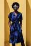 Person in blue and black patterned Sampa Dress with V-neckline and tie waist, standing straight against yellow wall.