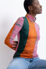 Colorful Stricta Turtleneck, large bold pink/orange/green stripes, blue jeans, white background, seated side pose.