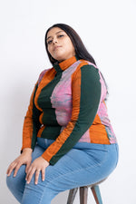 Multicolored Stricta Turtleneck in Carmine print, sitting on stool, bold orange, green, pink stripes, studio setting.
