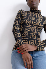 Person in high-neck top with black base and gold abstract patterns, hand on hip, against a white backdrop.