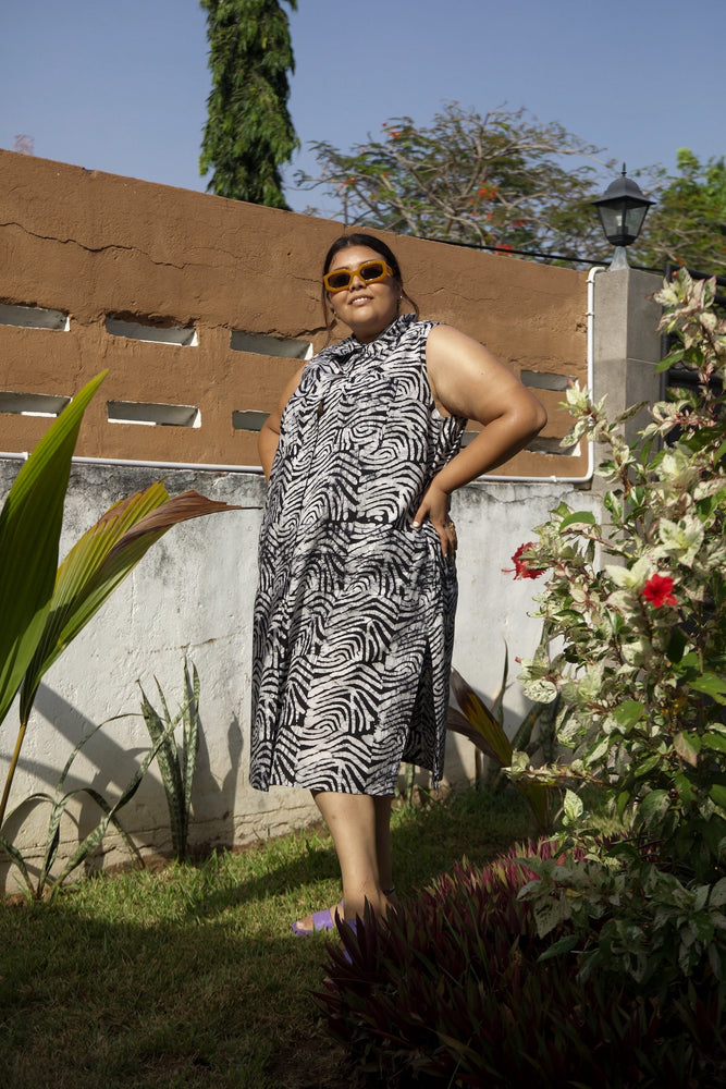 A woman wearing a zebra print dress stands in a garden posing beside blooming flowers and greenery under bright sunlight.