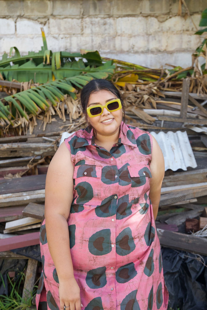 A woman in a pink patterned cotton shirtdress, standing in front of a pile of wood and greenery, wearing sunglasses.