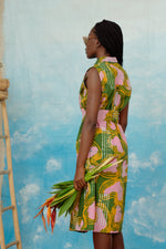 A person in a colorful batik print cotton dress holding flowers, standing against a painted sky backdrop.